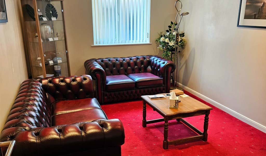 The inside of Edwin Pounds & Sons Funeral Directors Bradford branch arranging room. There is a red carpet with dark brown sofas.